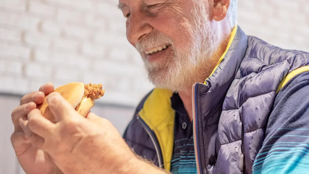 Un homme âgé barbu est assis tout en mangeant heureusement un sandwich au poulet dans un restaurant fast-food.