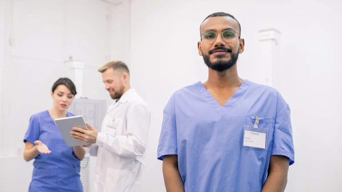 Jeune stagiaire métisse barbu debout devant la caméra sur fond de deux collègues à l'aide du pavé tactile