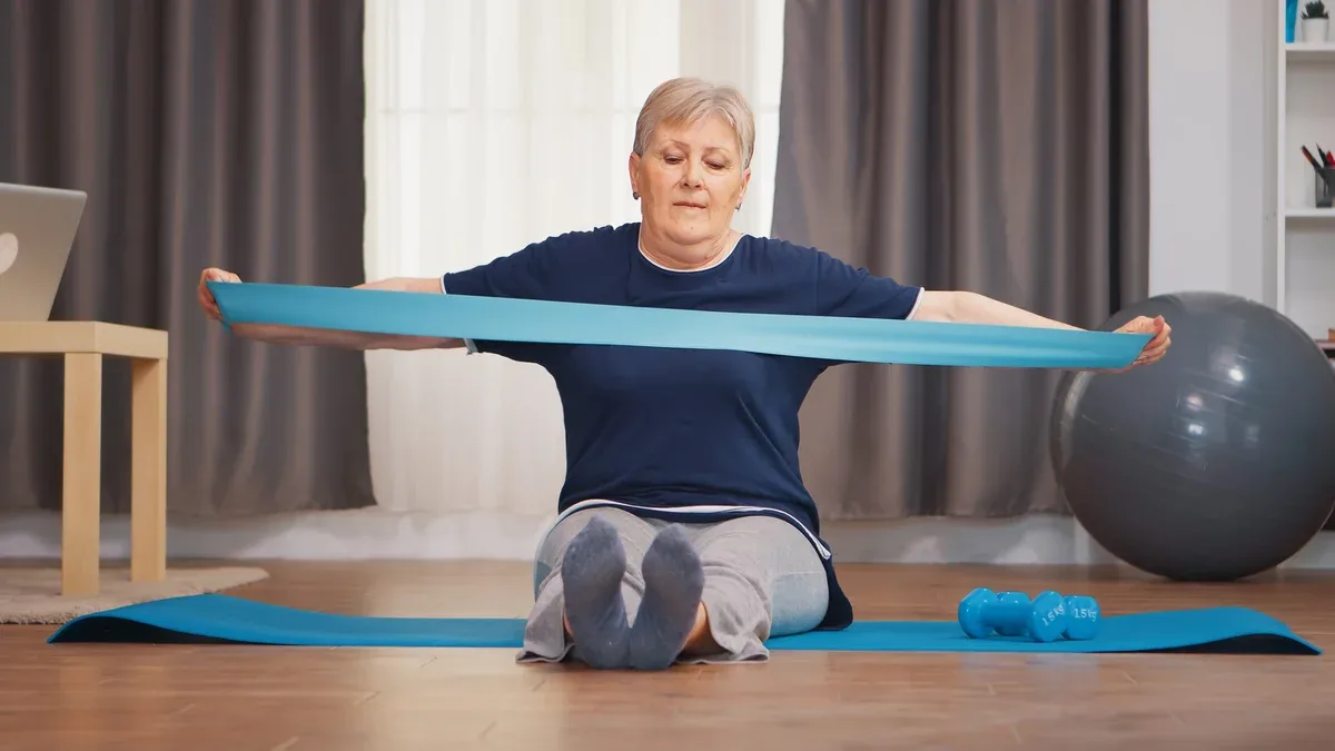 Senior woman formation avec bande de résistance assis sur un tapis de yoga. Vieille femme retraitée étirant la forme physique vivant un mode de vie sain à la retraite, s'entraînant à la maison