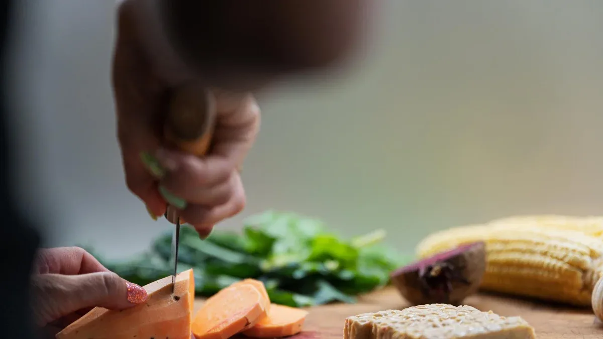 Vue en angle bas d'une femme coupant des patates douces sur une planche à découper pleine de légumes et de protéines de tempeh